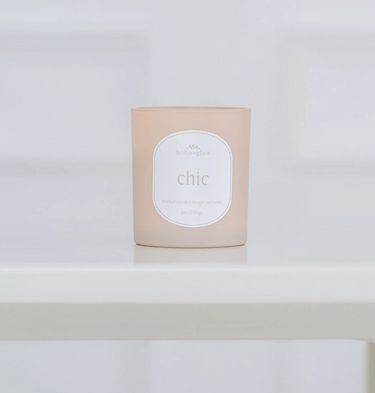 A frosted, light pinkish-beige candle in a glass jar sits on a white surface. The label reads